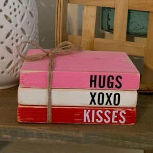 Puede incluir: Tres bloques de madera apilados pintados en rosa, blanco y rojo. El bloque superior dice "HUGS", el bloque del medio dice "XOXO" y el bloque inferior dice "KISSES".