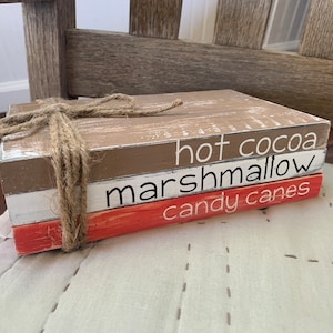 May include: Three stacked wooden blocks with the words "hot cocoa", "marshmallow", and "candy canes" painted in white on them. The blocks are tied together with twine.