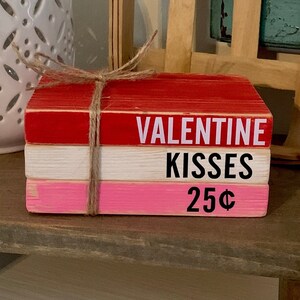 Puede incluir: Una pila de tres bloques de madera pintados en rojo, blanco y rosa. Los bloques están atados con un cordel. El bloque superior dice "VALENTINE KISSES 25¢".