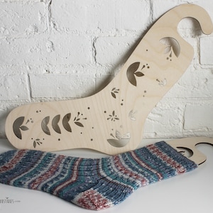 Peut inclure: Un outil de mise en forme de chaussettes en bois avec un motif floral. L'outil a un crochet sur le dessus pour l'accrocher. Une chaussette tricotée est affichée sur l'outil.