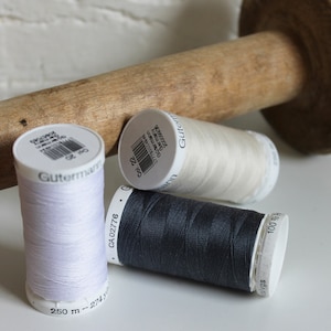 May include: Three spools of thread, one white, one off-white, and one dark grey, with the Gutermann brand name on the labels. The spools are sitting on a white surface next to a wooden spool.