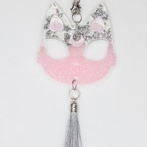 May include: A pink and silver glitter cat charm with a silver tassel. The charm is shaped like a cat's face with large eyes and a small nose. The charm is attached to a silver chain.