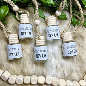 May include: Five small glass bottles with wooden tops and hanging cords. Each bottle has a label that reads "SONS AND RAE NEW CAR." The cords are made of a natural fiber and have wooden beads. The background is a mix of green leaves and a light-coloured, furry surface.