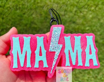 Könnte beinhalten: Ein rosa Lufterfrischer mit dem Wort "MAMA" in türkisfarbenen Buchstaben und einem türkisfarbenen Glitzerblitz. Der Lufterfrischer hat eine schwarze Schnur zum Aufhängen. Der Hintergrund ist verschwommenes grünes Gras.