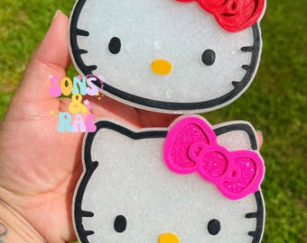 May include: Two Hello Kitty-shaped car air fresheners. The white faces have black outlines, black eyes, and yellow noses. One has a red bow, the other a pink glitter bow. The text "Sons & Ras" is visible.