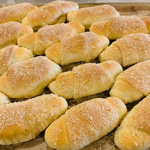 May include: A tray of freshly baked, golden-brown bread rolls, dusted with a light coating of crumbs. The rolls are crescent-shaped and arranged on a baking sheet, suggesting a homemade or bakery-fresh quality. The image evokes warmth and the appeal of baked goods.