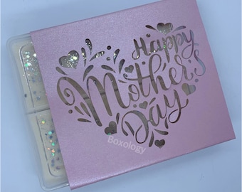 May include: A pink box with a heart-shaped cutout that says "Happy Mother's Day" in a decorative font. The box is filled with small, clear wax melts with glitter.