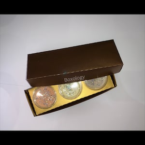 Pode incluir: Uma caixa marrom com a palavra "Boxology" impressa na parte superior. A caixa está aberta e contém três pequenos frascos redondos com conteúdo brilhante. Os frascos estão dispostos em uma fila sobre um fundo dourado.