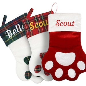 May include: Three personalized Christmas stockings with paw print designs. The stockings are red, white, and green with plaid trim. The names "Scout", "Scout", and "Belle" are embroidered on the stockings.