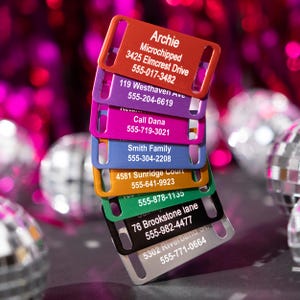 May include: A stack of colorful pet ID tags in various colors, including red, purple, pink, blue, orange, green, and silver. Each tag has a rectangular shape with rounded corners and includes text with a pet's name, address, and phone number.