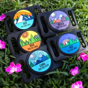 May include: A collection of black pet collar attachments with colorful circular designs featuring mountain and nature scenes. Each tag has a name and phone number. The tags are on a bed of green grass with pink flowers.