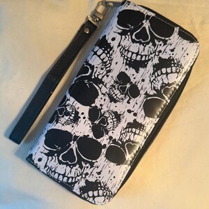 May include: A rectangular zip-around wallet featuring a repeating black and white graphic print of skulls wearing sunglasses. The wallet includes a detachable black wrist strap attached to a silver-toned metal clasp on the top edge of the wallet.