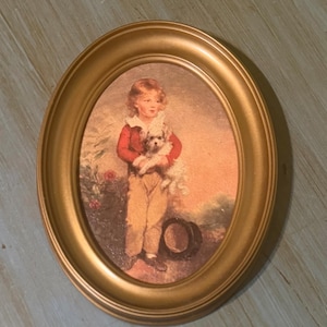 May include: Oval gold-toned framed print featuring a young person in a red jacket and tan pants, holding a small white dog. The print has a vintage aesthetic, with a hat on the ground and a soft, painterly style. The frame is a warm, metallic gold.