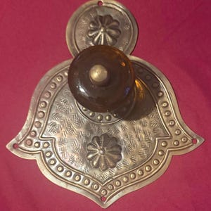 May include: A brass door plate with a bell-shaped design featuring embossed floral motifs, geometric patterns, and a border of small circles. A round, amber-colored glass knob with a brass center is mounted in the middle of the metal plate.