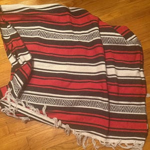 May include: A woven blanket featuring a repeating pattern of horizontal stripes in red, black, and white. The textile includes sections with a geometric zigzag design and is finished with a fringe of white threads along the bottom edge.
