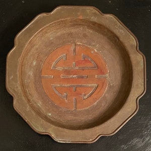 May include: A round brass tray with a scalloped edge and a weathered patina. The center features a circular inlay of reddish-brown material with a raised, metallic Shou symbol representing longevity, centered within the basin of the tray.