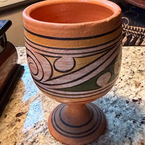 May include: A handmade terracotta goblet with a wide rim and a pedestal base. The goblet features a painted design with black, yellow, and green geometric patterns. The interior is a warm orange color.