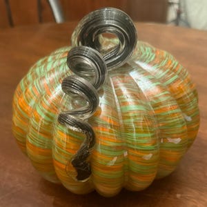 May include: A decorative glass pumpkin featuring a swirled pattern of orange and light green colors. The pumpkin has a ribbed, rounded body and a prominent, clear glass stem that curls into a spiral shape at the top of the object.