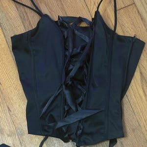 May include: A black sleeveless corset top featuring thin spaghetti straps and a central vertical lace-up closure with matching black ribbon ties. The garment is constructed with structured seams and includes a side zipper for fastening.