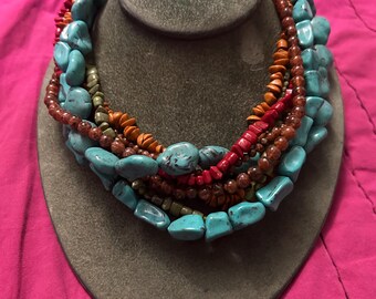 May include: Multi-strand necklace with turquoise, red, orange, and brown beads. The necklace is layered and has a chunky, bohemian style.
