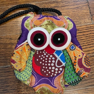 May include: A colorful owl-shaped fabric purse with a black drawstring closure. The owl has large white eyes with black pupils and red eyelids. The purse is made of patchwork fabric with various patterns and colors, including purple, green, red, and gold.