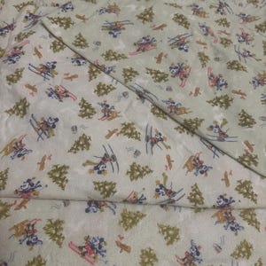 May include: A light green fabric featuring a repeating pattern of Mickey Mouse figures skiing and sledding, interspersed with dark green pine trees and small brown wooden signposts. The material has a soft, textured appearance throughout.