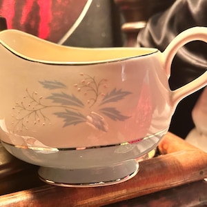 May include: A vintage cream pitcher with a light blue base and a floral design. The pitcher has a curved handle and a spout for easy pouring. The rim is trimmed with gold. The floral design features blue leaves and small gold-colored dots.