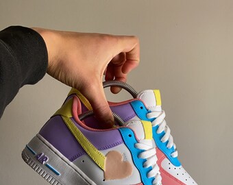 May include: White low-top sneakers with a pink, purple, blue, and yellow color block design. A pink heart is painted on the side of the shoe.
