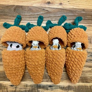 May include: Four handmade plush carrots with green tops and orange bodies. Each carrot has a small, stuffed animal peeking out. The stuffed animals are gray and white. The carrots are arranged on a wooden surface.
