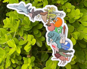 Könnte beinhalten: Ein bunter Aufkleber mit einem Design zum Thema Florida Keys. Der Aufkleber zeigt eine Vielzahl von Tieren und Pflanzen, darunter ein Seekuh, ein Flamingo, ein Pelikan, eine Palme, ein Fisch, eine Fledermaus, ein Flugzeug, eine Muschel, eine Krabbe und ein Alligator. Der Aufkleber enthält auch den Text "Evacuation Route".
