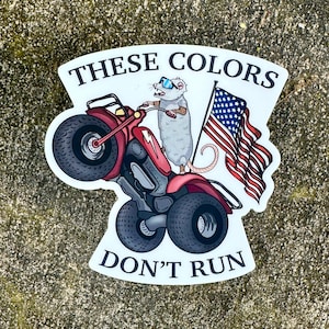 May include: A white sticker with a red, white, and blue American flag, a gray possum wearing sunglasses, and a red mini motorcycle. The sticker says "These Colors Don't Run".