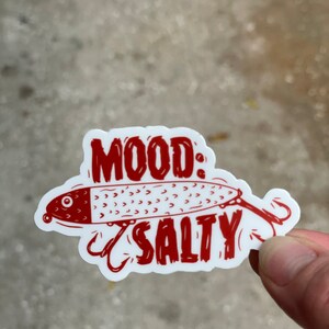 以下が含まれることがあります： 赤い白のステッカーに、アニメーションの釣りルアーと「MOOD: SALTY」の文字が書かれています。