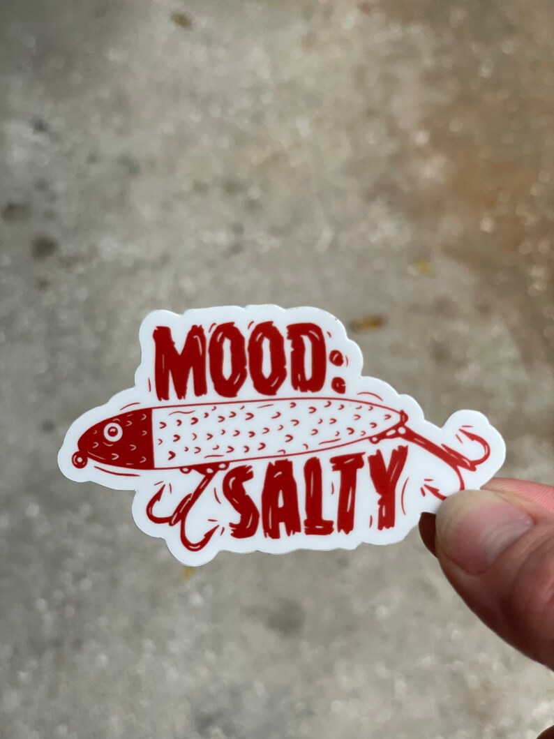 以下が含まれることがあります： 赤い白のステッカーに、アニメーションの釣りルアーと「MOOD: SALTY」の文字が書かれています。