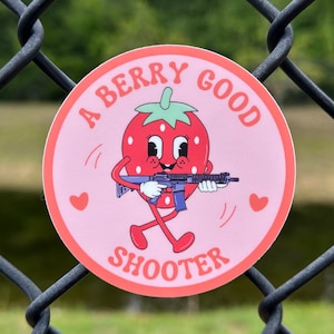 May include: A pink and red sticker with a cartoon strawberry holding a rifle. The sticker says "A Berry Good Shooter" in a retro font.
