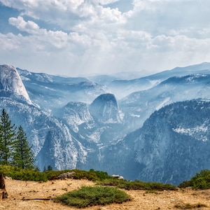 Puede incluir: Una vista panorámica de una cordillera con una gran formación rocosa en la distancia. Las montañas están cubiertas de una neblina azul claro y el cielo es azul brillante con nubes blancas.