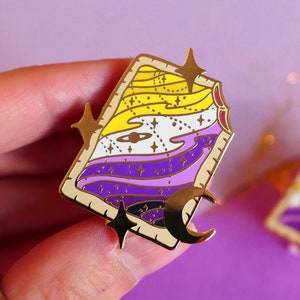 Peut inclure: Épinglette en émail doré avec un croissant de lune et des étoiles. L'épingle représente un drapeau de fierté non binaire stylisé avec des rayures jaunes, blanches, violettes et bleues.