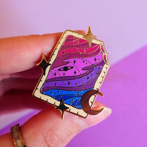 Pode incluir: Pin de esmalte dourado com uma imagem estilizada de uma paisagem celestial com uma lua crescente, estrelas e uma galáxia giratória em tons de rosa, roxo e azul.