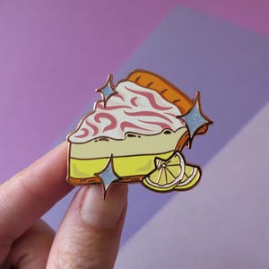 May include: A gold-tone enamel pin of a slice of lemon meringue pie with a swirl of pink meringue and a lemon wedge on top. The pin has a sparkly finish and is perfect for adding a touch of whimsy to your clothing or bag.