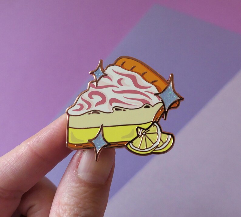 May include: A gold-tone enamel pin of a slice of lemon meringue pie with a swirl of pink meringue and a lemon wedge on top. The pin has a sparkly finish and is perfect for adding a touch of whimsy to your clothing or bag.
