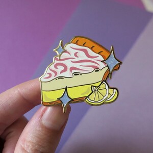 May include: A gold-tone enamel pin of a slice of lemon meringue pie with pink swirls and a lemon wedge. The pin has a sparkly blue glitter effect.