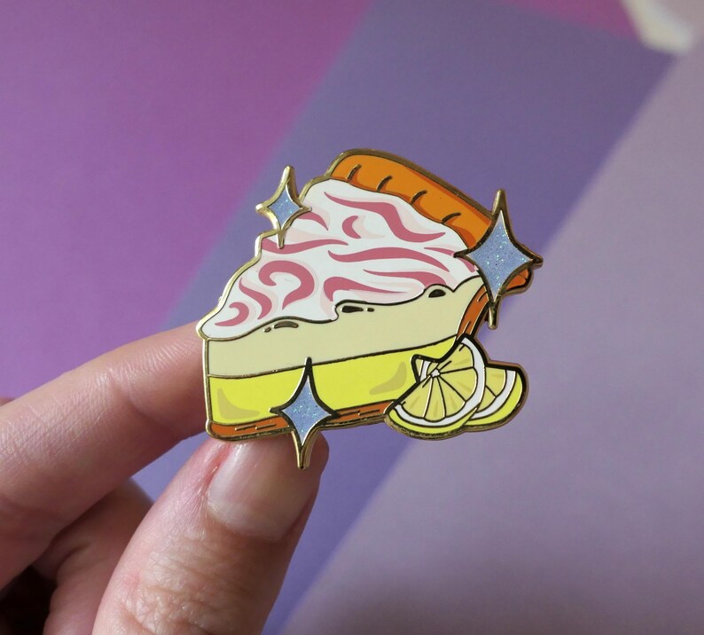 May include: A gold-tone enamel pin of a slice of lemon meringue pie with pink swirls and a lemon wedge. The pin has a sparkly blue glitter effect.
