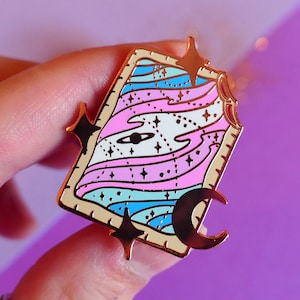 May include: A gold-colored enamel pin with a rectangular design featuring a galaxy-themed illustration in pink, blue, and white. The pin includes a planet, stars, and a crescent moon, with black star accents on the edges.