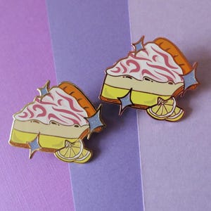 May include: Two enamel pins shaped like slices of lemon meringue pie with pink swirls and a yellow lemon wedge. The pins have a gold finish and glitter accents.
