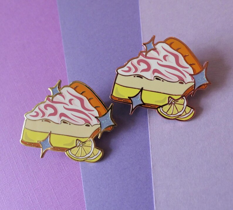 May include: Two enamel pins shaped like slices of lemon meringue pie with pink swirls and a yellow lemon wedge. The pins have a gold finish and glitter accents.