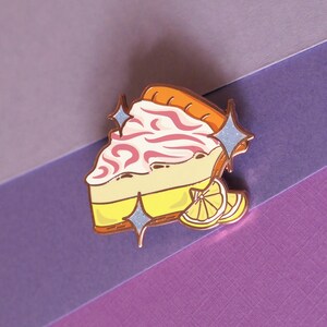 May include: A gold-tone enamel pin of a slice of lemon meringue pie with pink swirls and a lemon wedge. The pin has a sparkly finish and is perfect for adding a touch of whimsy to your clothing or bag.