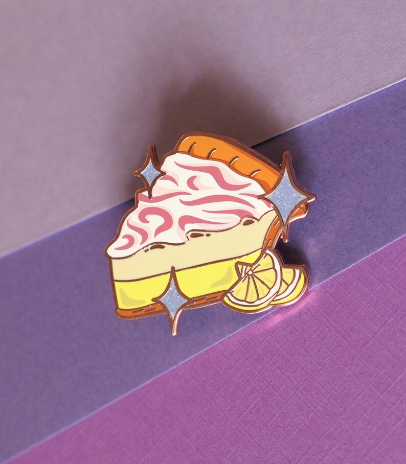 May include: A gold-tone enamel pin of a slice of lemon meringue pie with pink swirls and a lemon wedge. The pin has a sparkly finish and is perfect for adding a touch of whimsy to your clothing or bag.