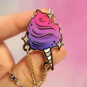 Puede incluir: Un pin de esmalte dorado en forma de remolino de algodón de azúcar con un efecto degradado de rosa a morado. El pin tiene una cadena unida a la parte inferior y presenta detalles de purpurina.