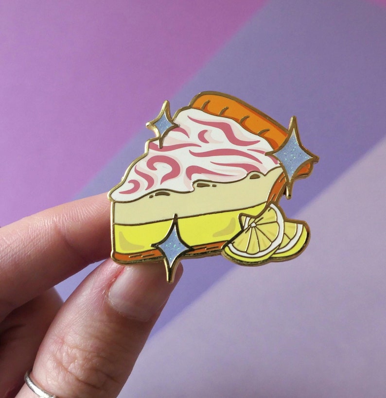 May include: A gold-tone enamel pin featuring a slice of lemon meringue pie with pink swirls and a lemon wedge. The pin has a sparkly finish.