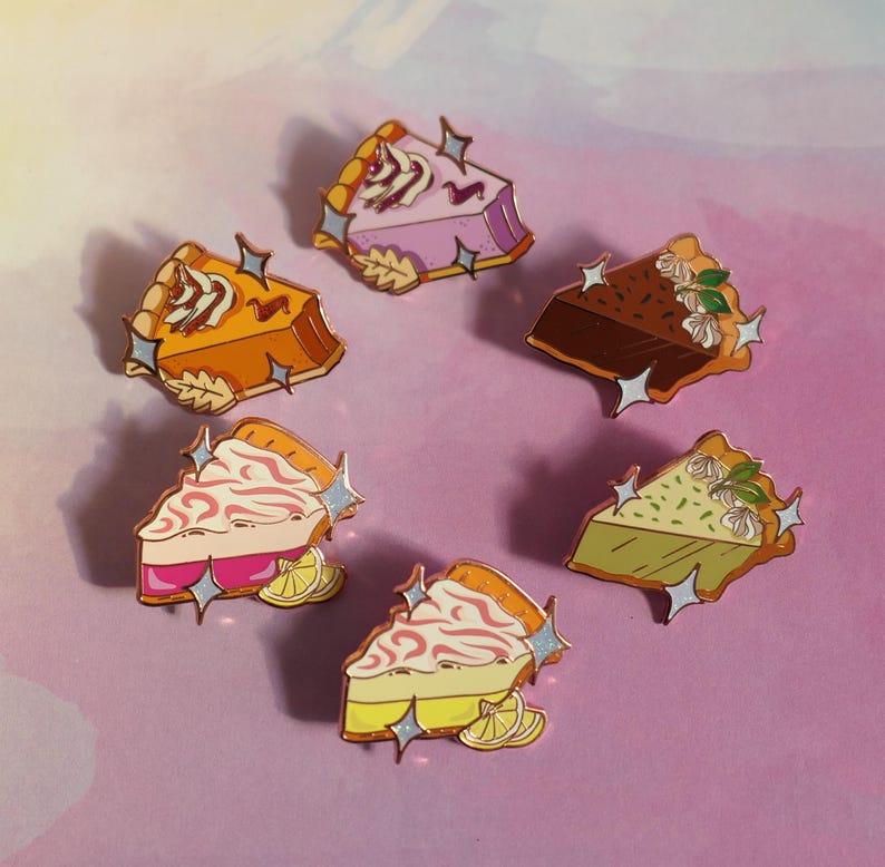 May include: Six enamel pins shaped like slices of pie with different fillings and crusts. The pins are on a pink and purple background. The fillings include pumpkin, chocolate, lemon, and berry.