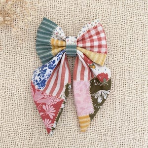 May include: A fabric hair bow featuring a patchwork design with various patterns including red and white gingham, green stripes, floral prints, and solid yellow sections. The bow is constructed from multiple textile segments stitched together.
